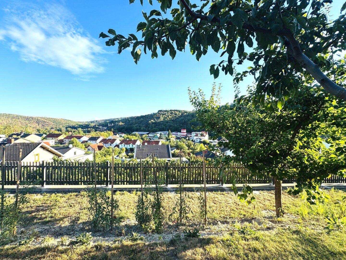 Weitläufiger Blick über eine ländliche Siedlung und bewaldete Hügel unter blauem Himmel.
