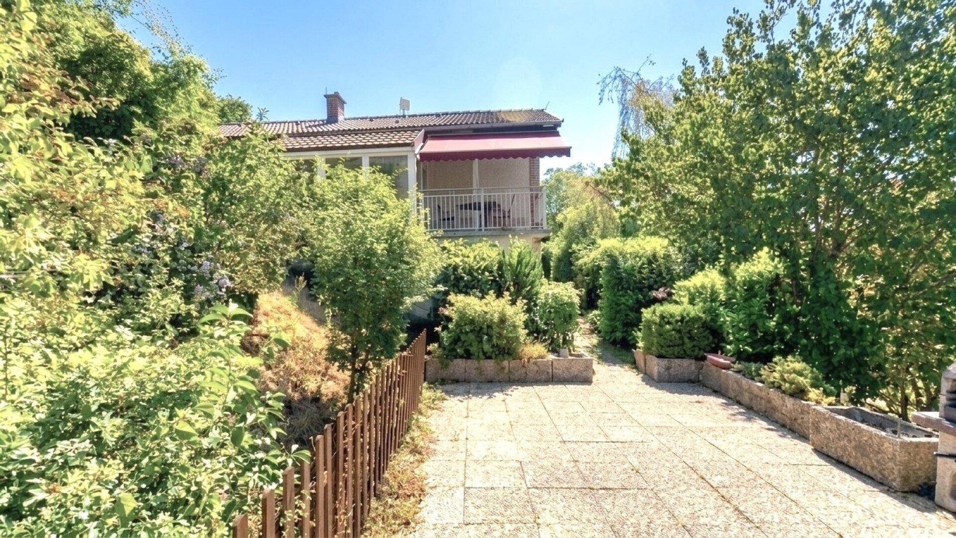 Gepflasterter Weg führt zum Haus, umgeben von gepflegten Büschen und Bäumen, mit einem Balkon im Hintergrund.