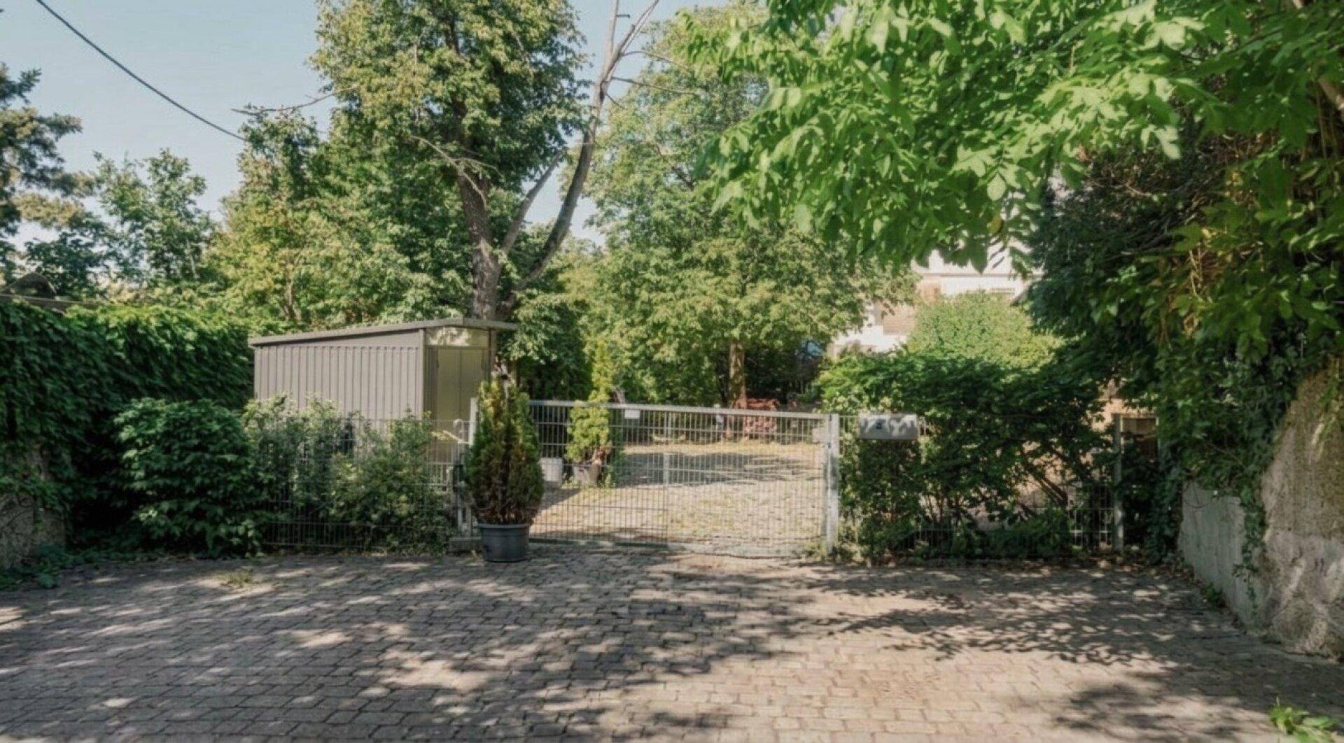 Gepflasterter Parkplatz mit Metalltor und Gartenschuppen, umgeben von Bäumen und Hecken.