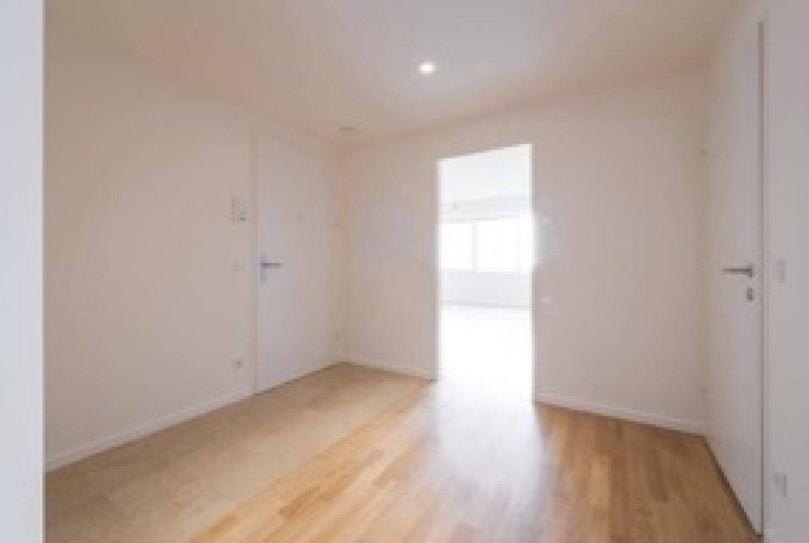 Leerer Raum mit Laminatboden und weiß gestrichenen Wänden, hell durch angrenzendes Zimmer.