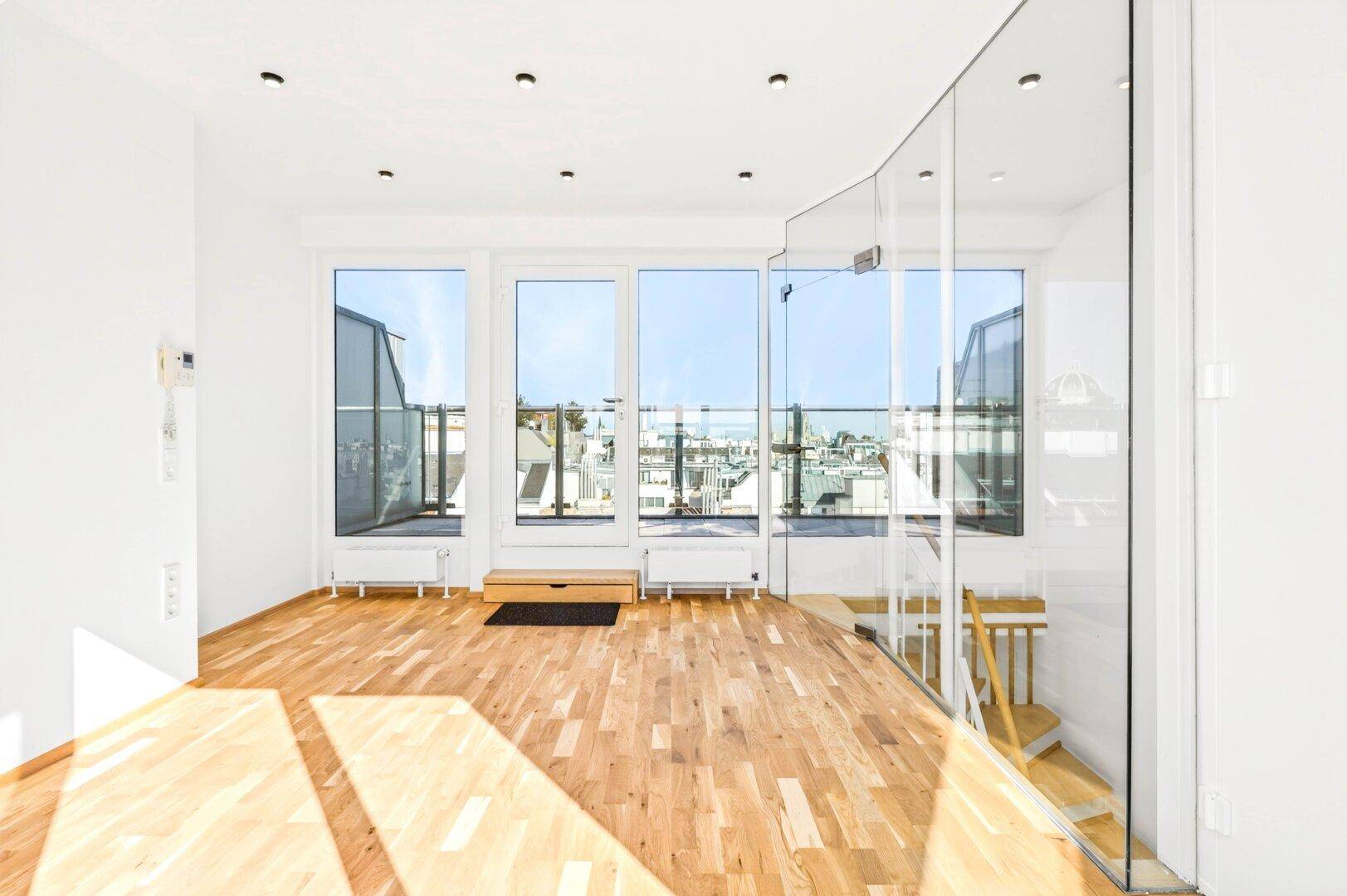 Heller, leerer Raum mit Holzboden, großen Fenstern und Zugang zu einem Balkon oder einer Terrasse.