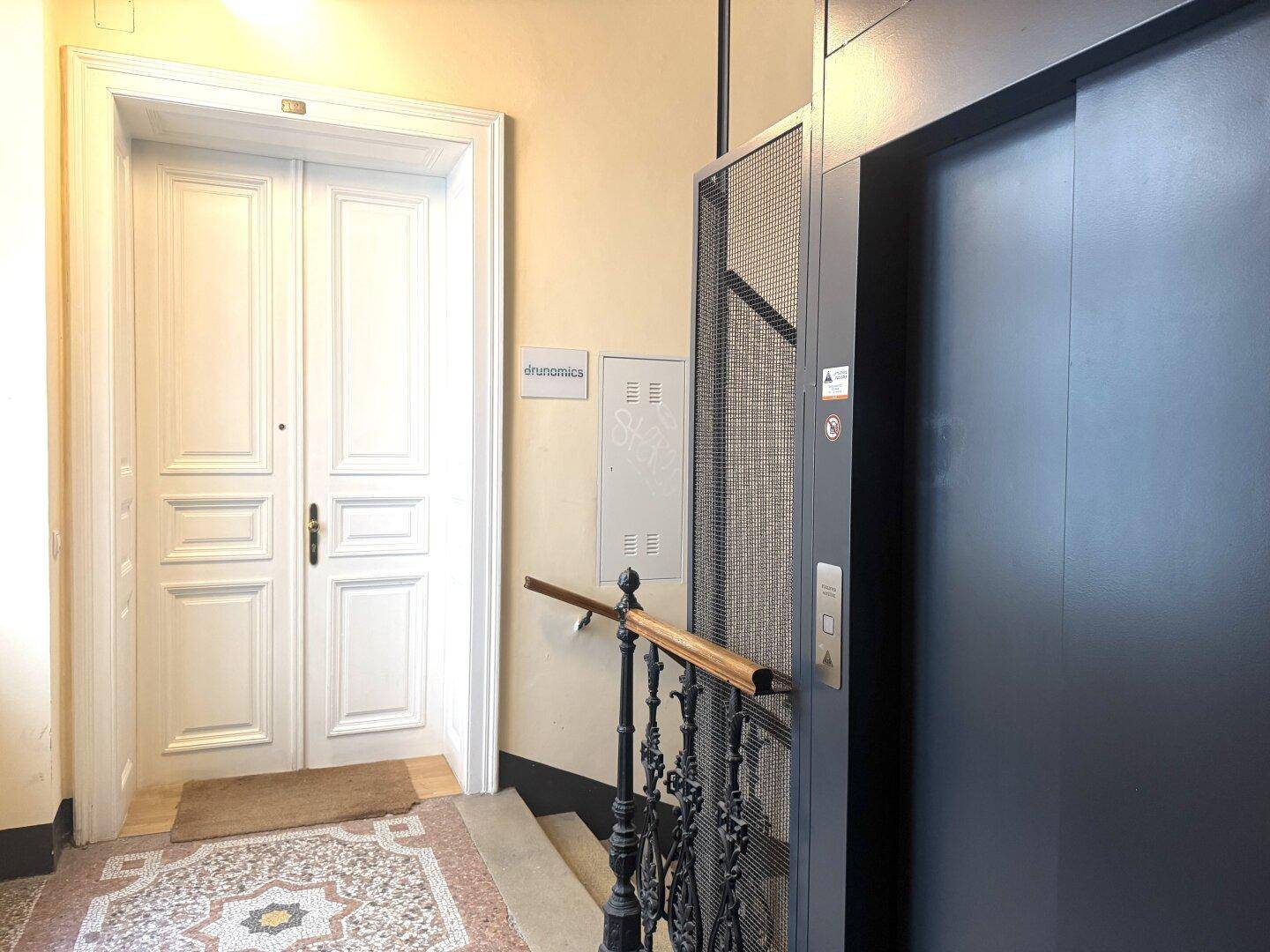 Eingangsbereich mit modernem Aufzug, Treppe und einer großen Doppeltür, die zur Wohnung führt.