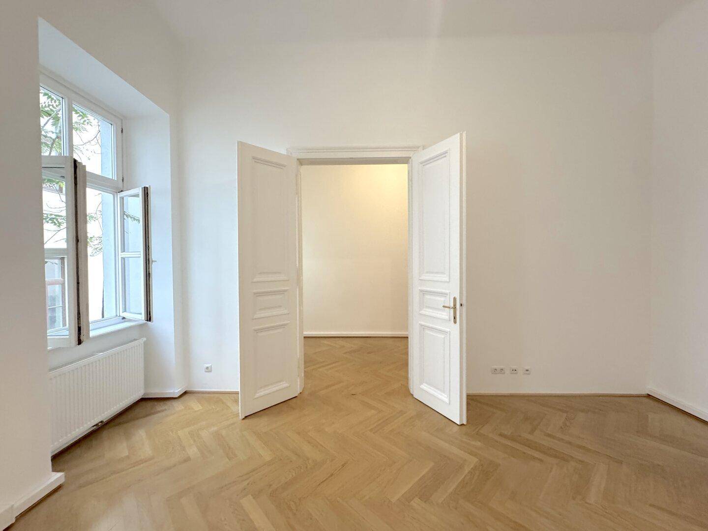 Großer, heller Raum mit Fischgrätparkett und hohen Doppeltüren, die zu einem weiteren Zimmer führen.