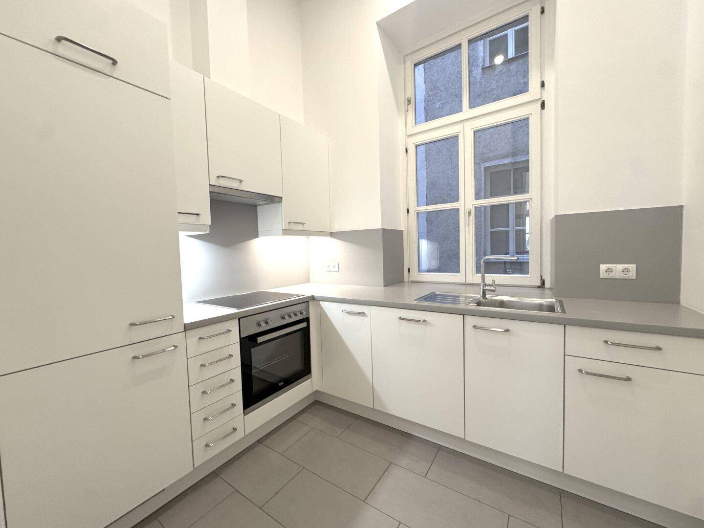 Moderne Einbauküche in Weiß und Grau mit integriertem Backofen und Spülbecken unter dem Fenster.