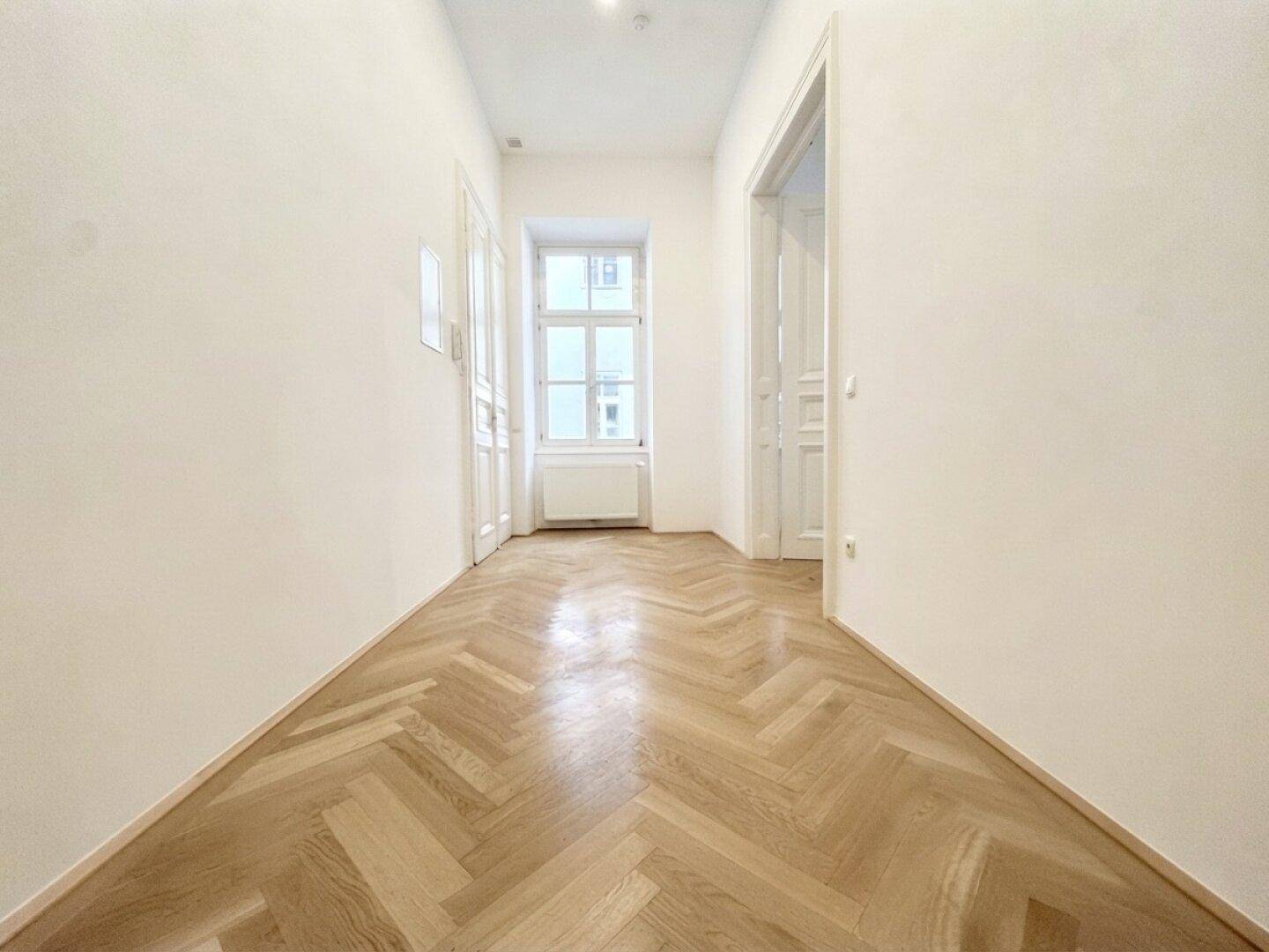 Langer, heller Flur mit Fischgrätparkett und einem Fenster am Ende, das viel Tageslicht spendet.