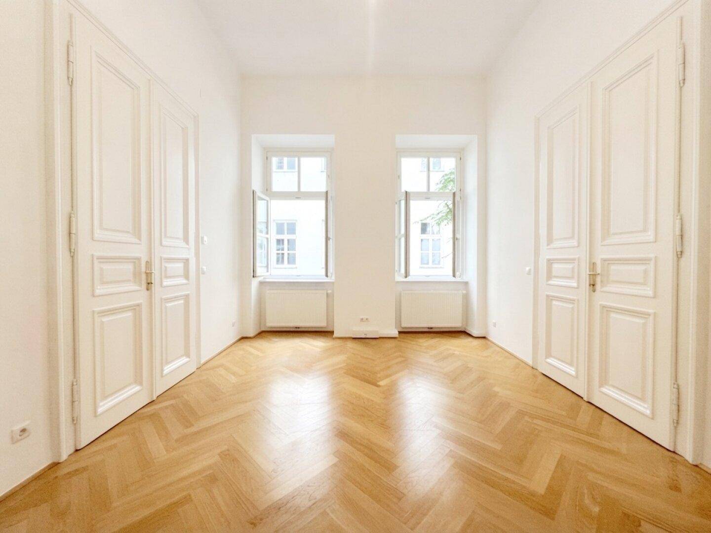 Geräumiger, leerer Raum mit klassischem Fischgrätparkett und zwei hohen Fenstern, die viel Licht hereinlassen.