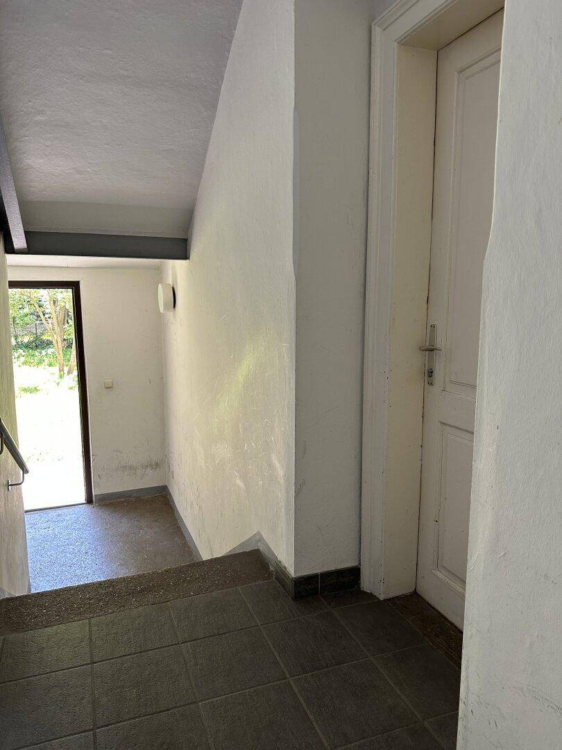 Heller Treppenaufgang mit weiß gestrichenen Wänden und dunklen Fliesen, führt zum Ausgang ins Grüne.