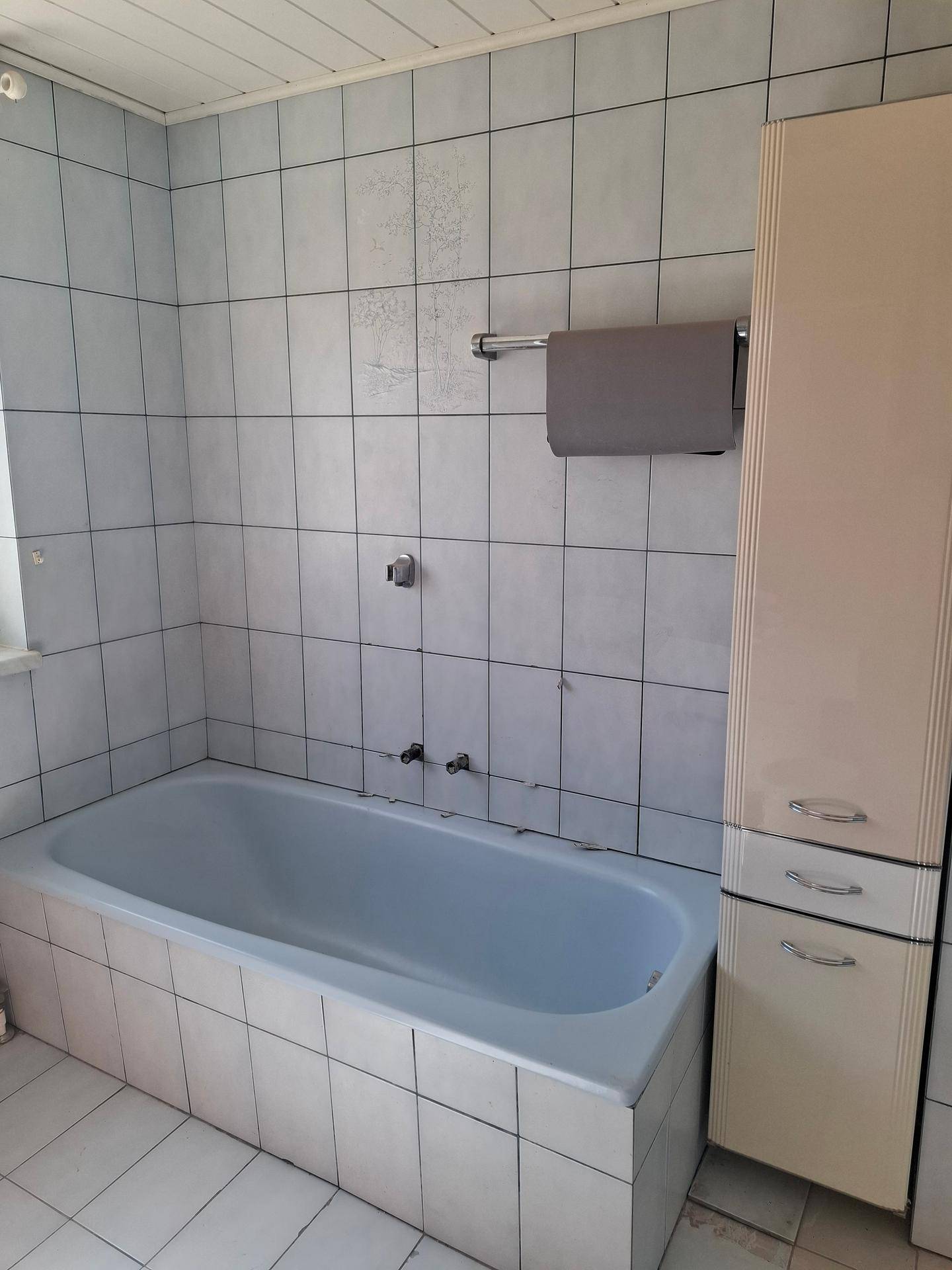 Badezimmer mit hellblauen Fliesen, Badewanne und einem schmalen Hochschrank in Standardausstattung.
