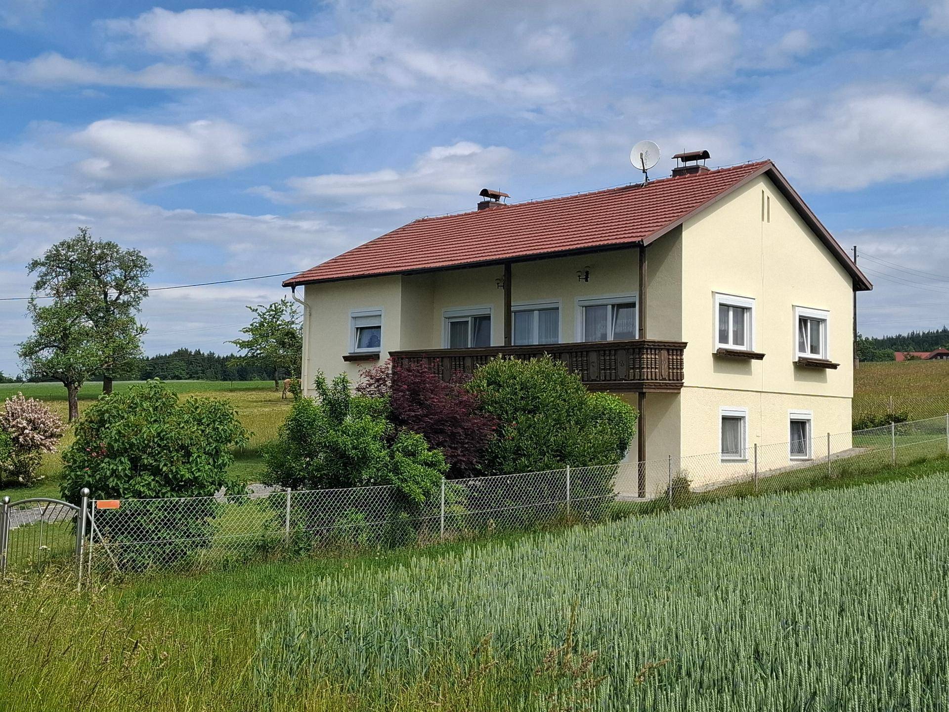 Hausfassade mit Balkon und rotem Ziegeldach, umgeben von grünen Feldern unter blauem Himmel.
