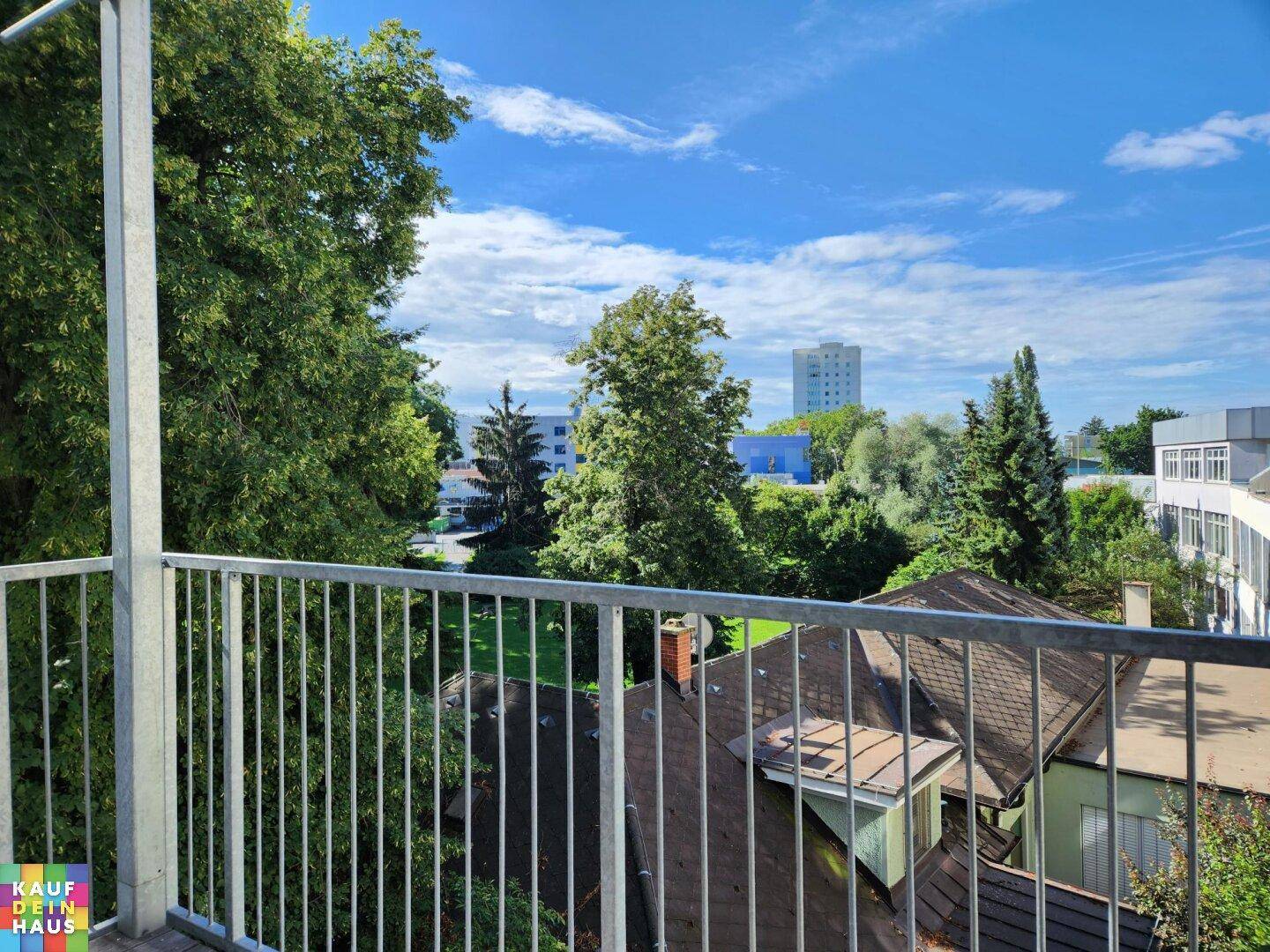 Blick vom Balkon ins Grüne mit Bäumen, Dächern und einem Gebäude in der Ferne.