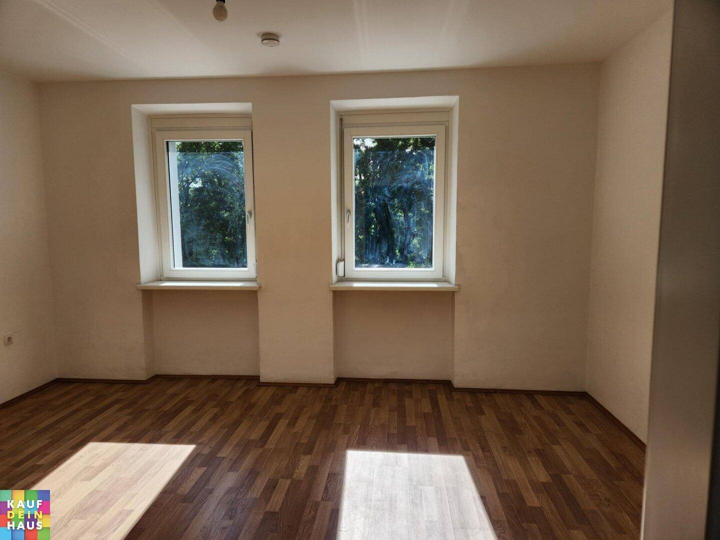 Leeres Zimmer mit Laminatboden und zwei Fenstern, die viel Tageslicht hereinlassen.