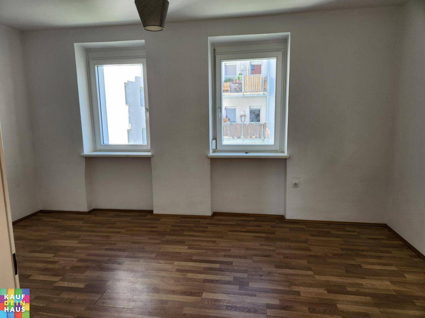 Helles Zimmer mit Laminatboden und Blick aus den Fenstern auf gegenüberliegende Balkone.