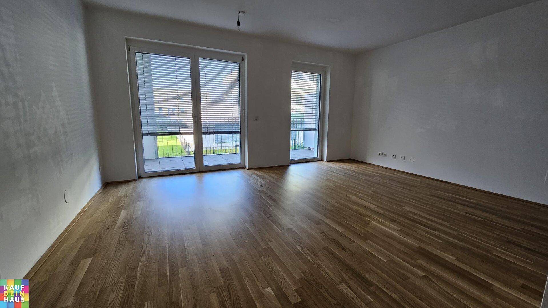 Helles, leeres Wohnzimmer mit Holzboden und großen Fenstern zum Außenbereich.