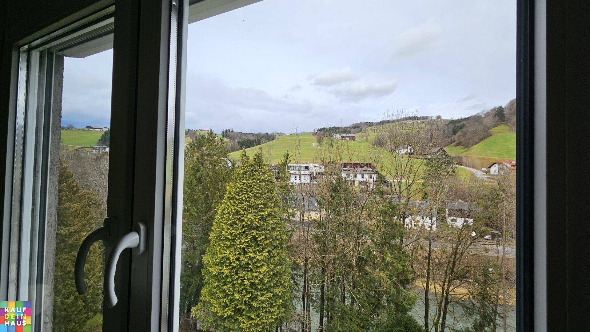 Malerischer Ausblick aus dem Fenster auf eine grüne Hügellandschaft mit Fluss und Häusern.