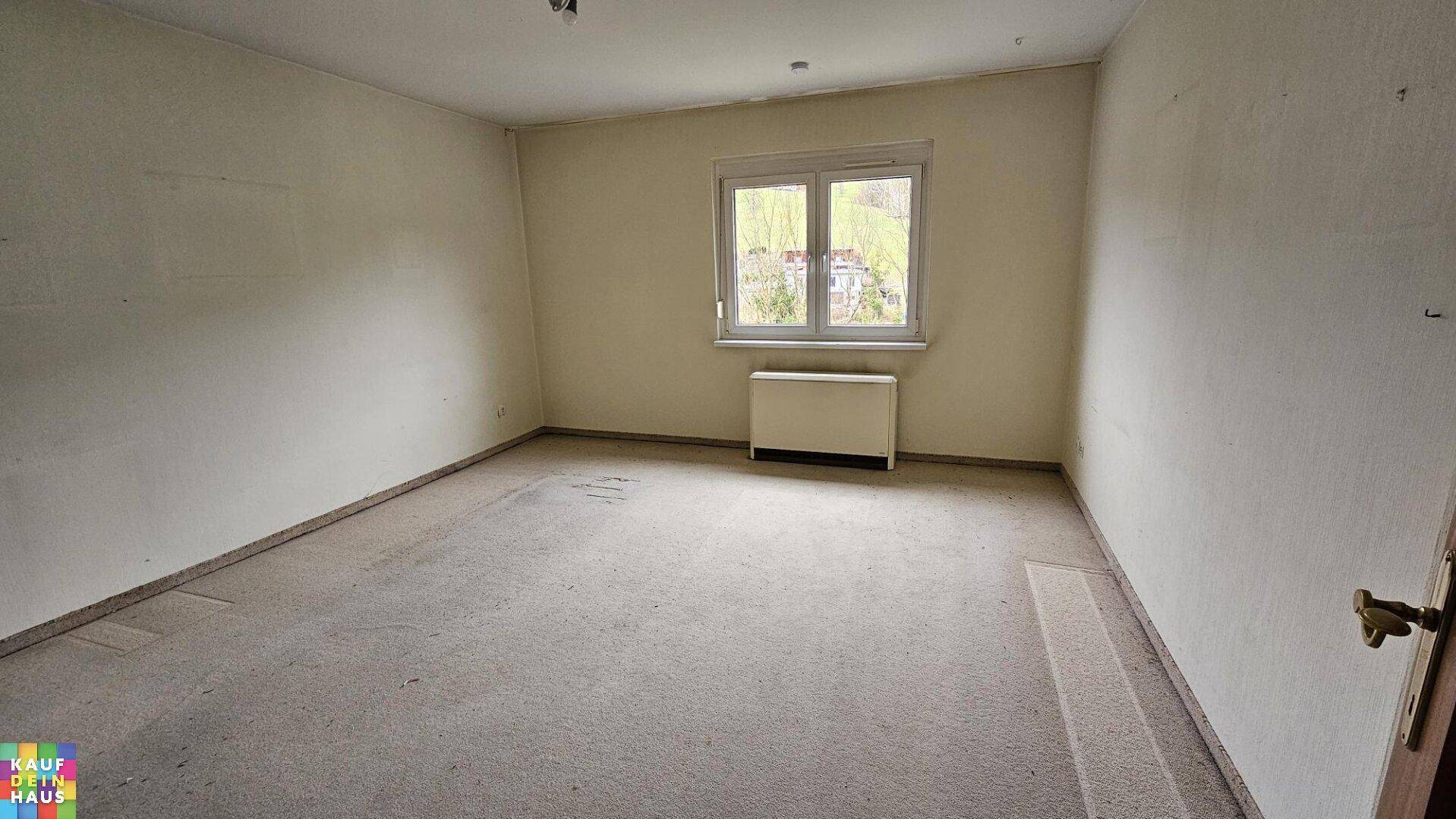 Helles Zimmer mit Teppichboden und Fenster, das einen ruhigen Blick ins Grüne bietet.