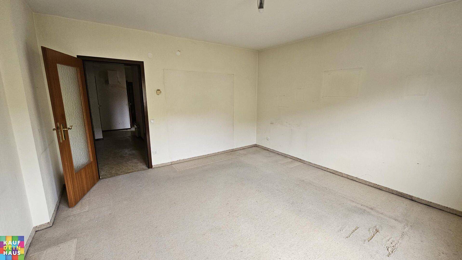 Leeres Zimmer mit hellem Teppichboden und offener Tür, das auf eine Modernisierung wartet.