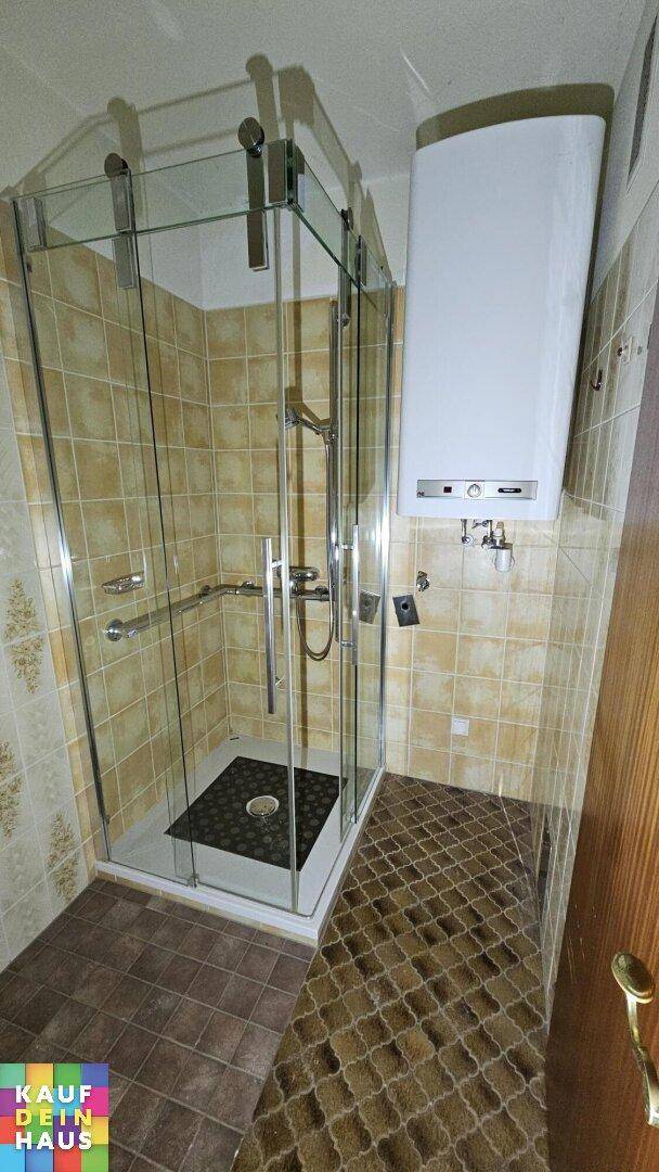 Badezimmer mit moderner Glasduschkabine, beigefarbenen Fliesen und Warmwasserbereiter an der Wand.