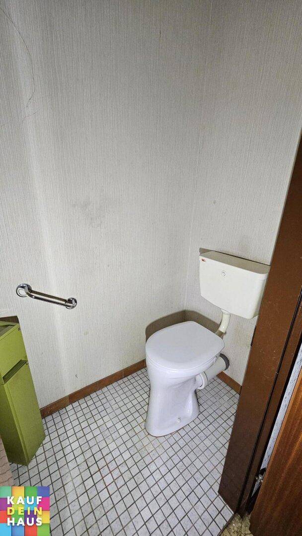 Kleines WC mit Toilette, Haltegriff und weiß gefliestem Boden, funktional und einfach gehalten.