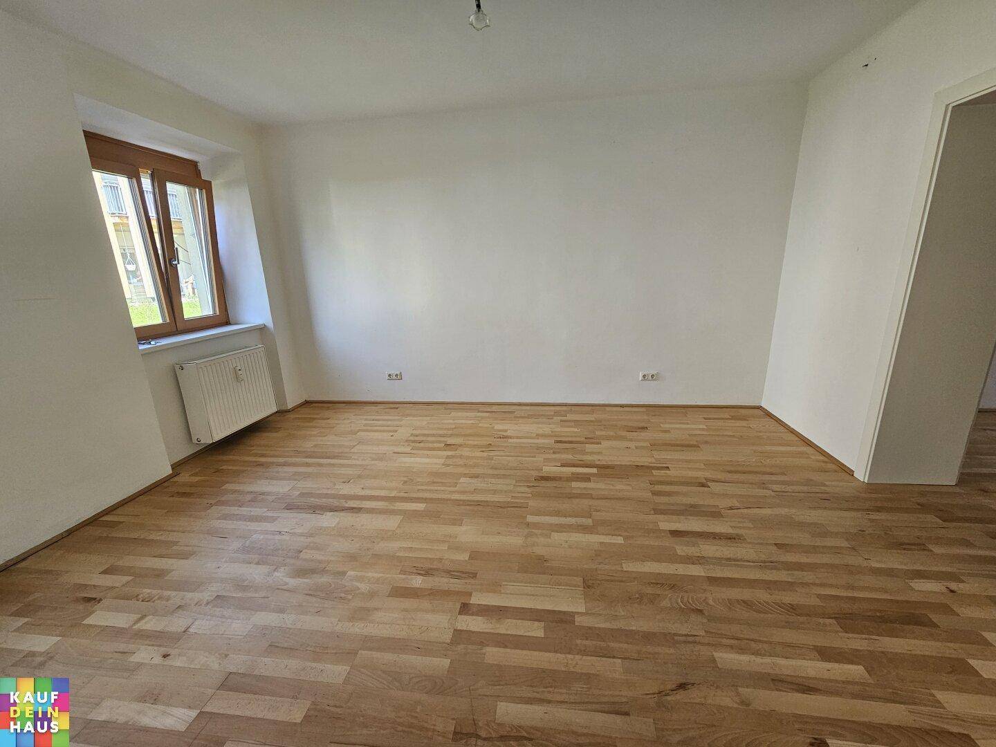 Geräumiger, leerer Raum mit Holzboden, einem Fenster und weiß gestrichenen Wänden.