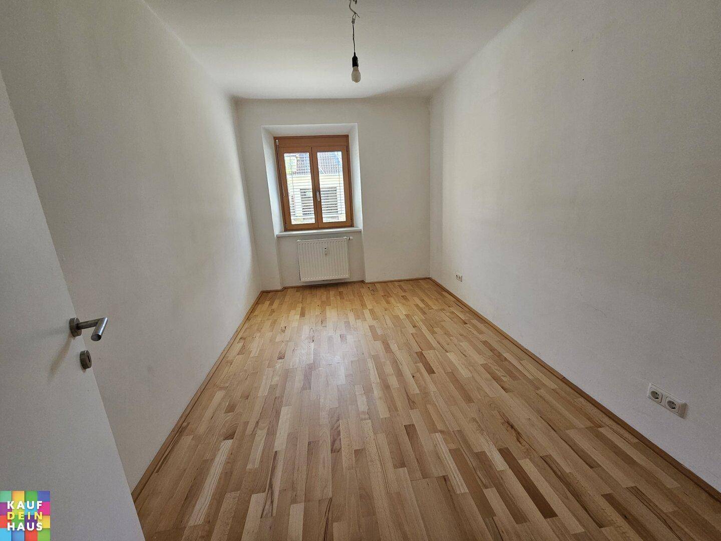 Leerer Raum mit Holzboden, einem Fenster und einer Tür, die zu einem anderen Bereich führt.