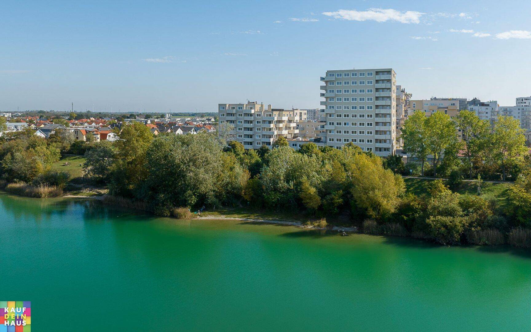 Weitläufiger Blick über einen ruhigen See mit Wohngebäuden und grüner Uferlandschaft im Hintergrund.