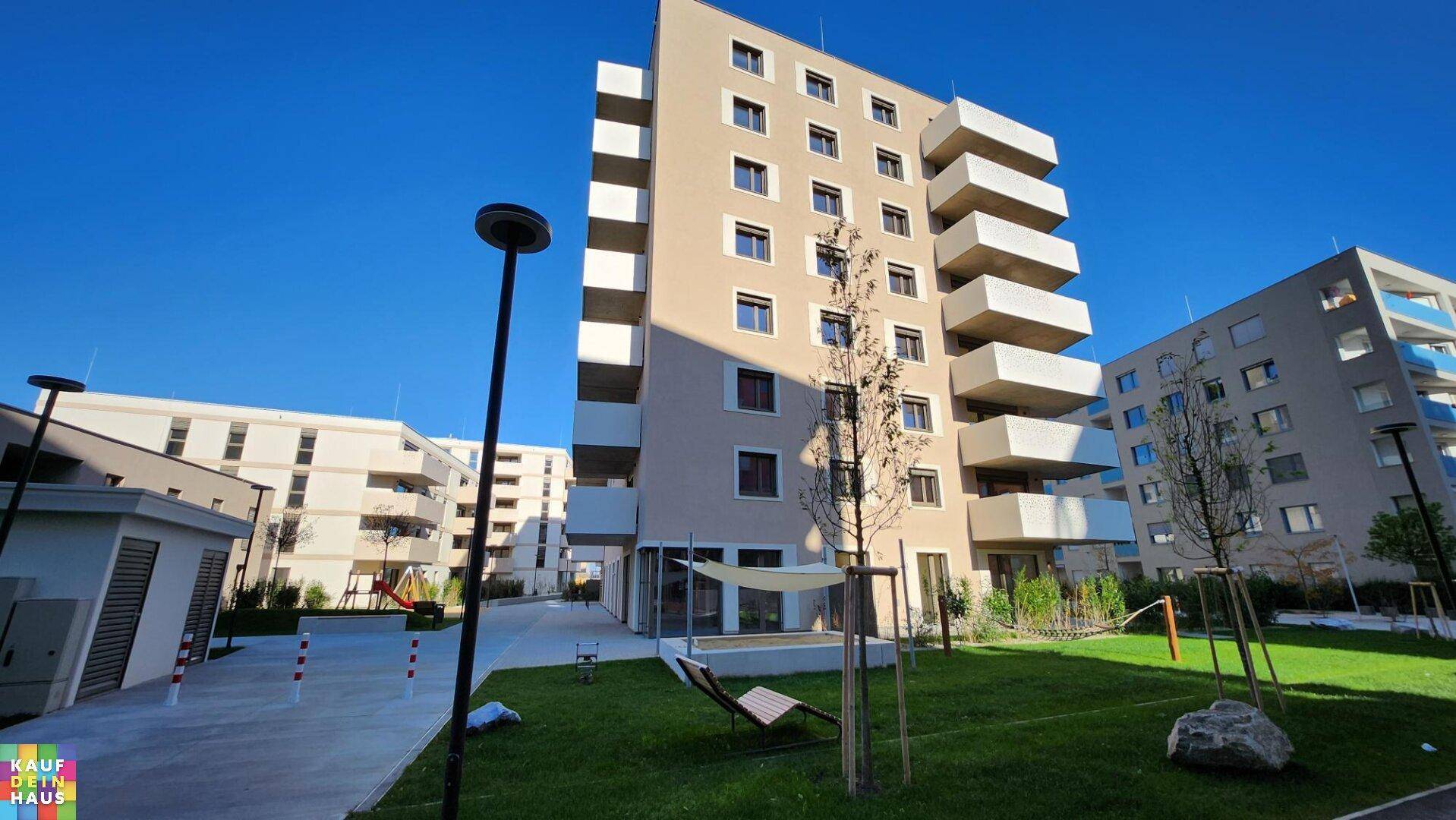 Weitläufige Außenanlage eines Neubau-Wohnkomplexes mit Grünflächen, Bäumen und einem Spielbereich.