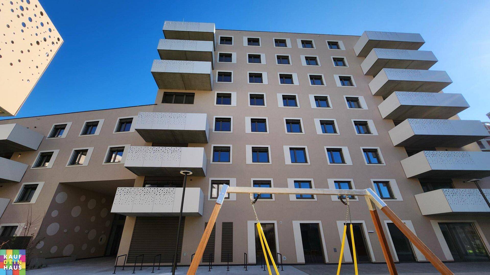Fassade eines modernen Wohngebäudes mit vielen Balkonen und einem Spielbereich im Vordergrund.