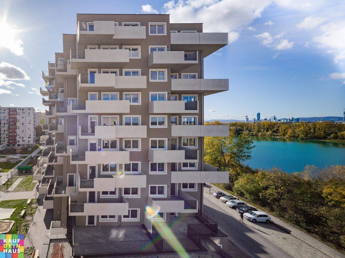 Modernes Wohngebäude mit vielen Balkonen und Seeblick unter strahlend blauem Himmel.