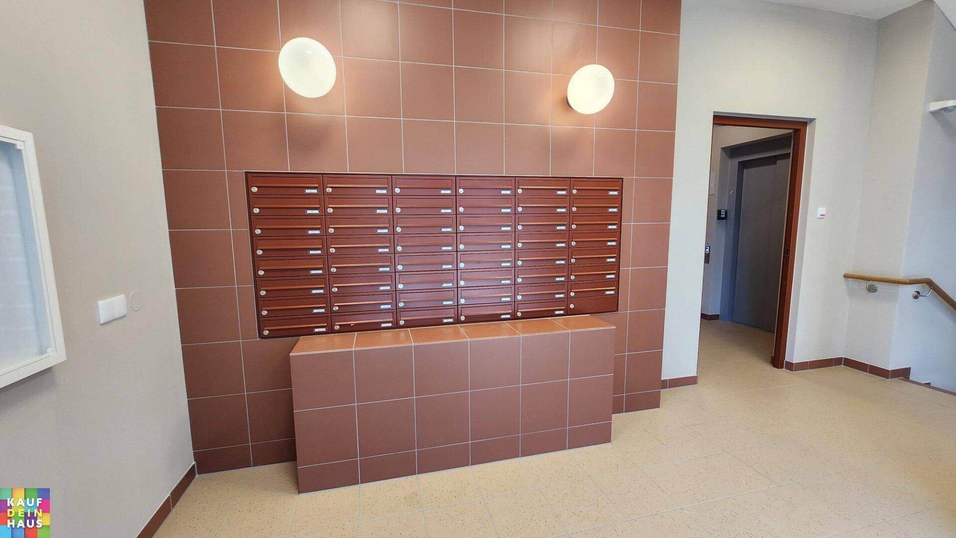 Eingangsbereich mit Briefkästen und braunen Fliesen an den Wänden, beleuchtet von Wandlampen.