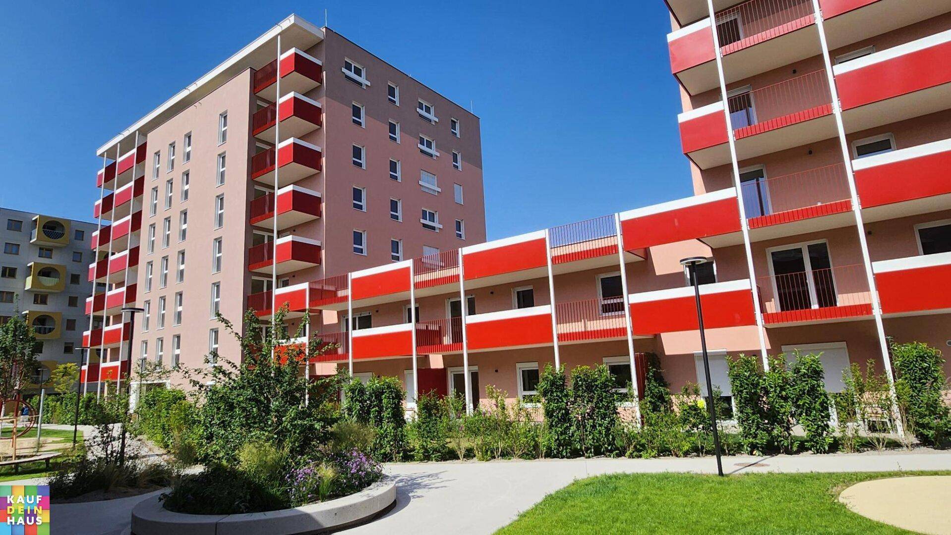 Moderne Wohngebäude mit roten Balkonen und gepflegten Grünflächen unter blauem Himmel.