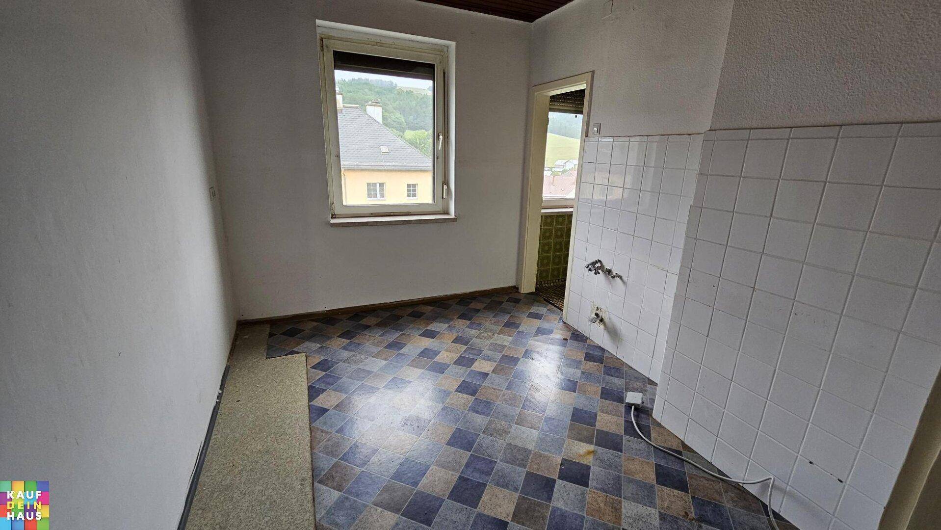 Leere Küche mit gefliesten Wänden und gemustertem Boden, mit Zugang zu einem weiteren Raum.