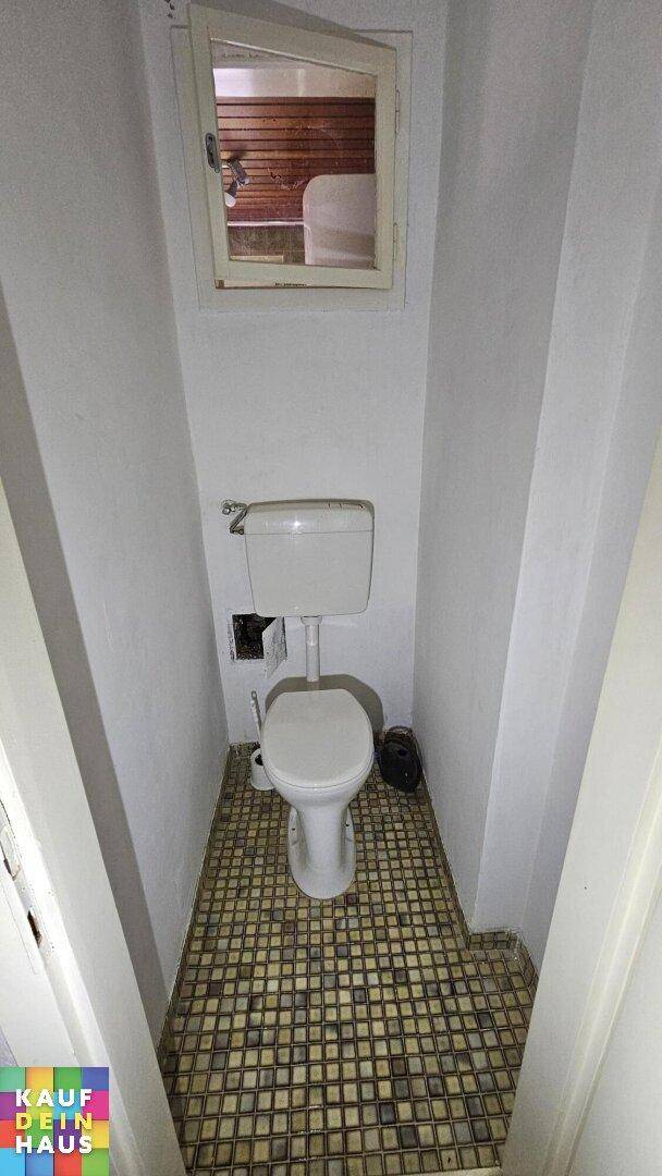 Separate Toilette mit kleinem Fenster und gemusterten Bodenfliesen, in einfachem Zustand.