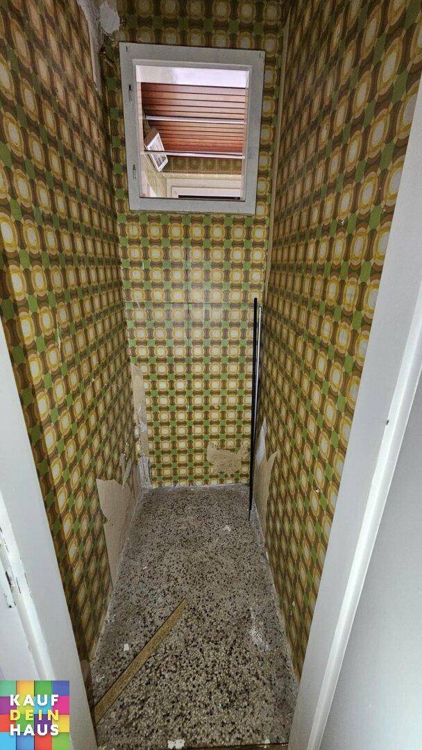 Schmaler Raum mit auffälliger gemusterter Tapete und Fenster, benötigt Renovierung.