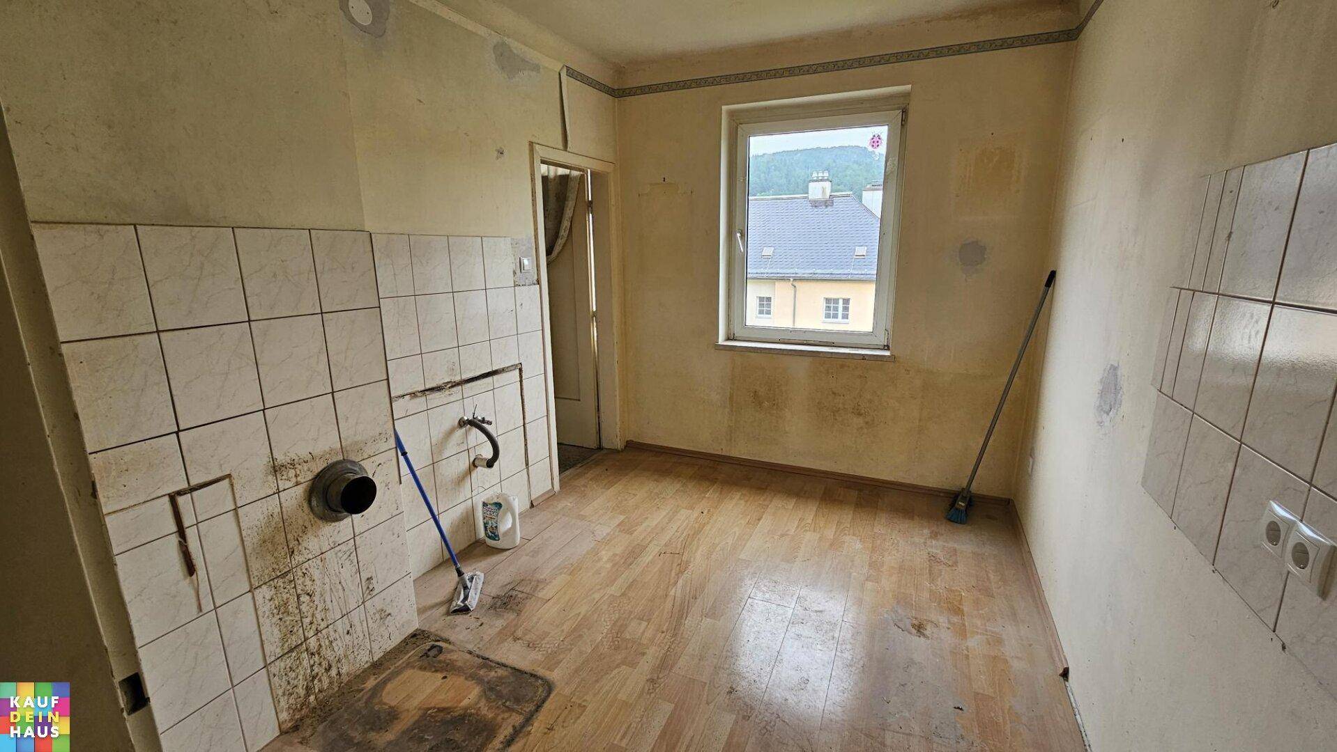 Leere Küche mit alten Wandfliesen, sichtbaren Anschlüssen und Holzdielenboden.