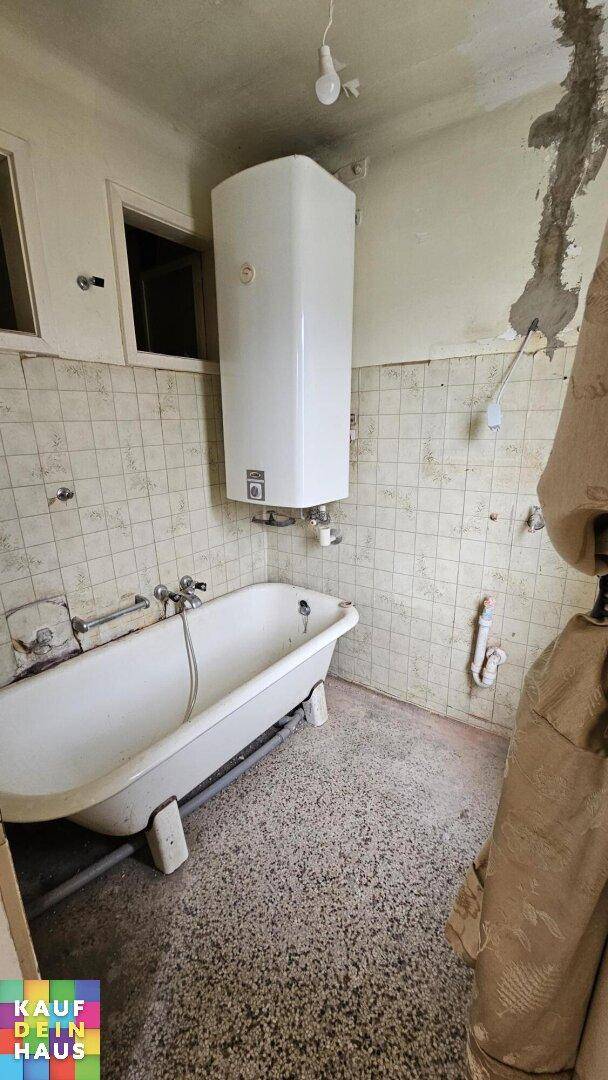 Badezimmer mit alter Badewanne, gefliesten Wänden und einem Warmwasserboiler.