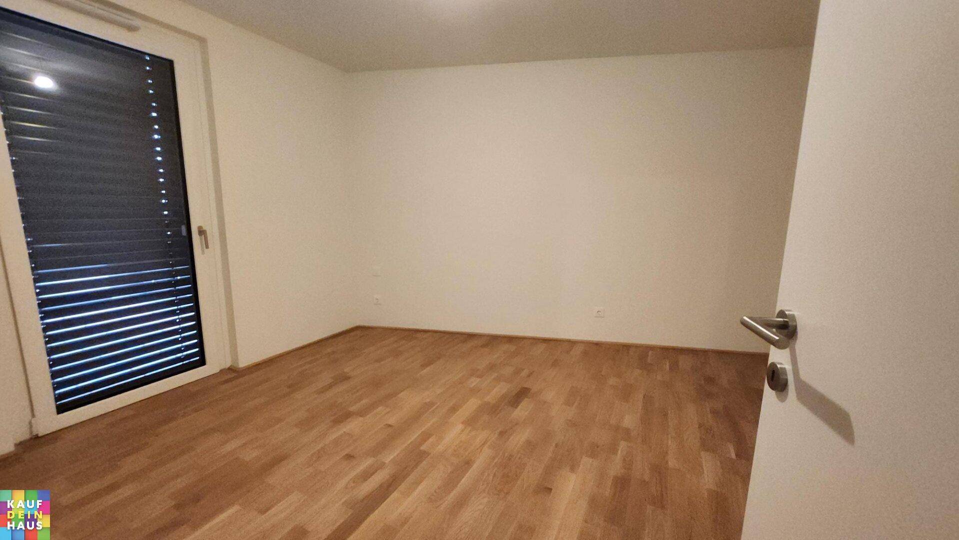 Leeres Schlafzimmer mit hellem Parkettboden, weißen Wänden und Fenster mit Jalousien.