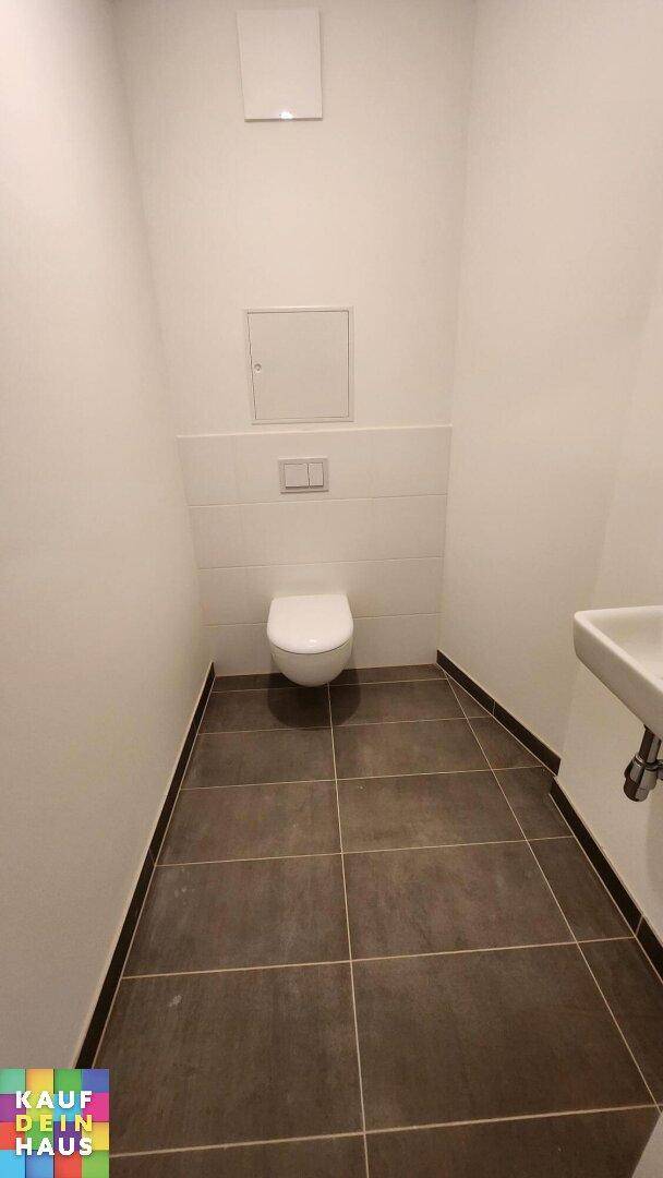 Separates Gäste-WC mit wandhängender Toilette, kleinem Waschbecken und dunklen Bodenfliesen.