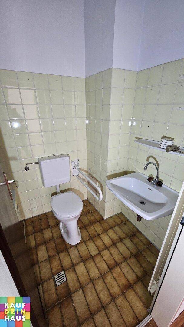 Badezimmer mit hellgrünen Fliesen, WC mit Haltegriffen und Waschbecken.