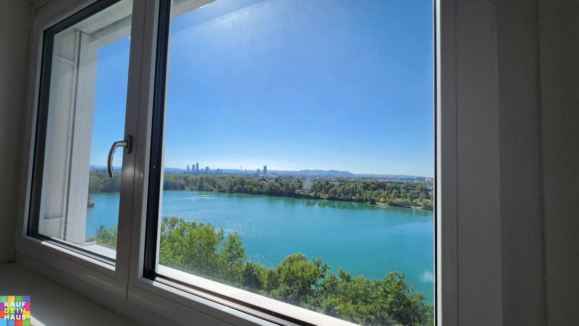 Fensterblick auf einen türkisfarbenen See mit Stadtkulisse und grünen Ufern an einem sonnigen Tag.