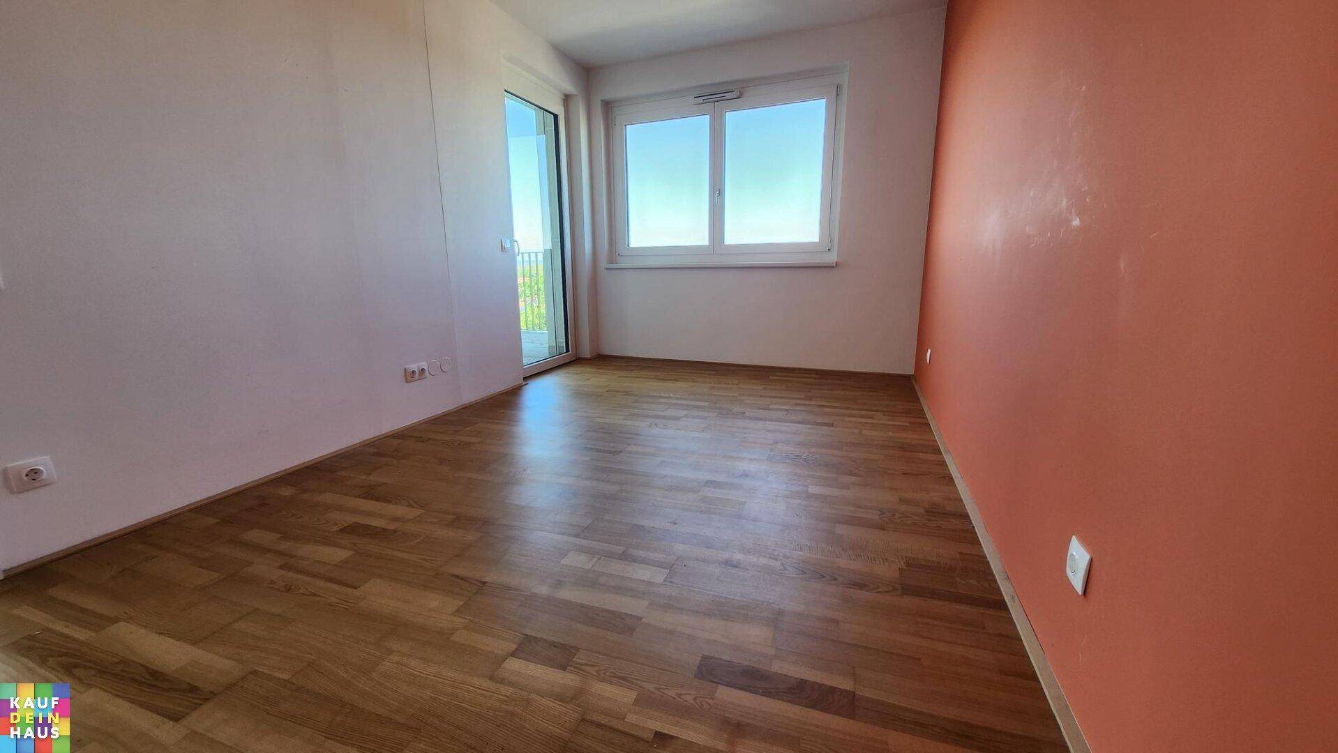 Helles Zimmer mit Parkettboden, einer farbigen Akzentwand und Zugang zu einem Balkon.