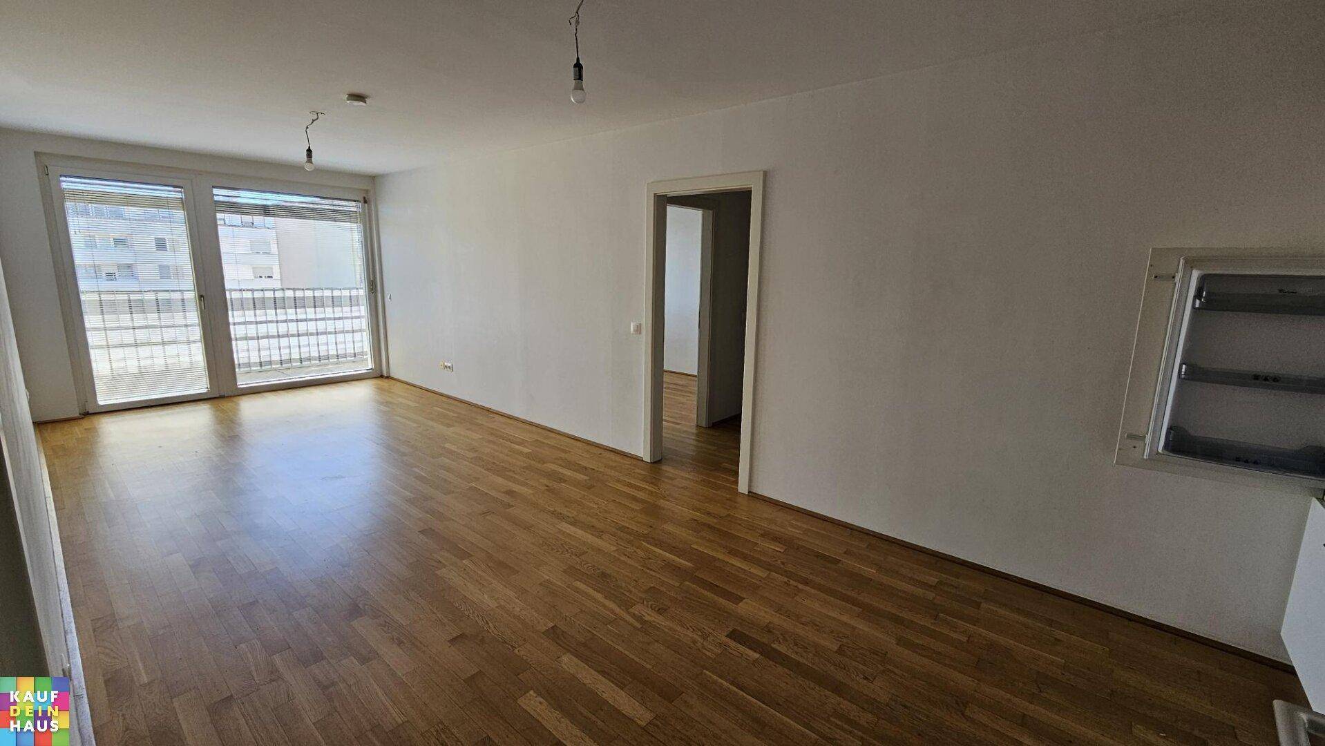 Helles, leeres Wohnzimmer mit Parkettboden und Zugang zu einem Balkon.