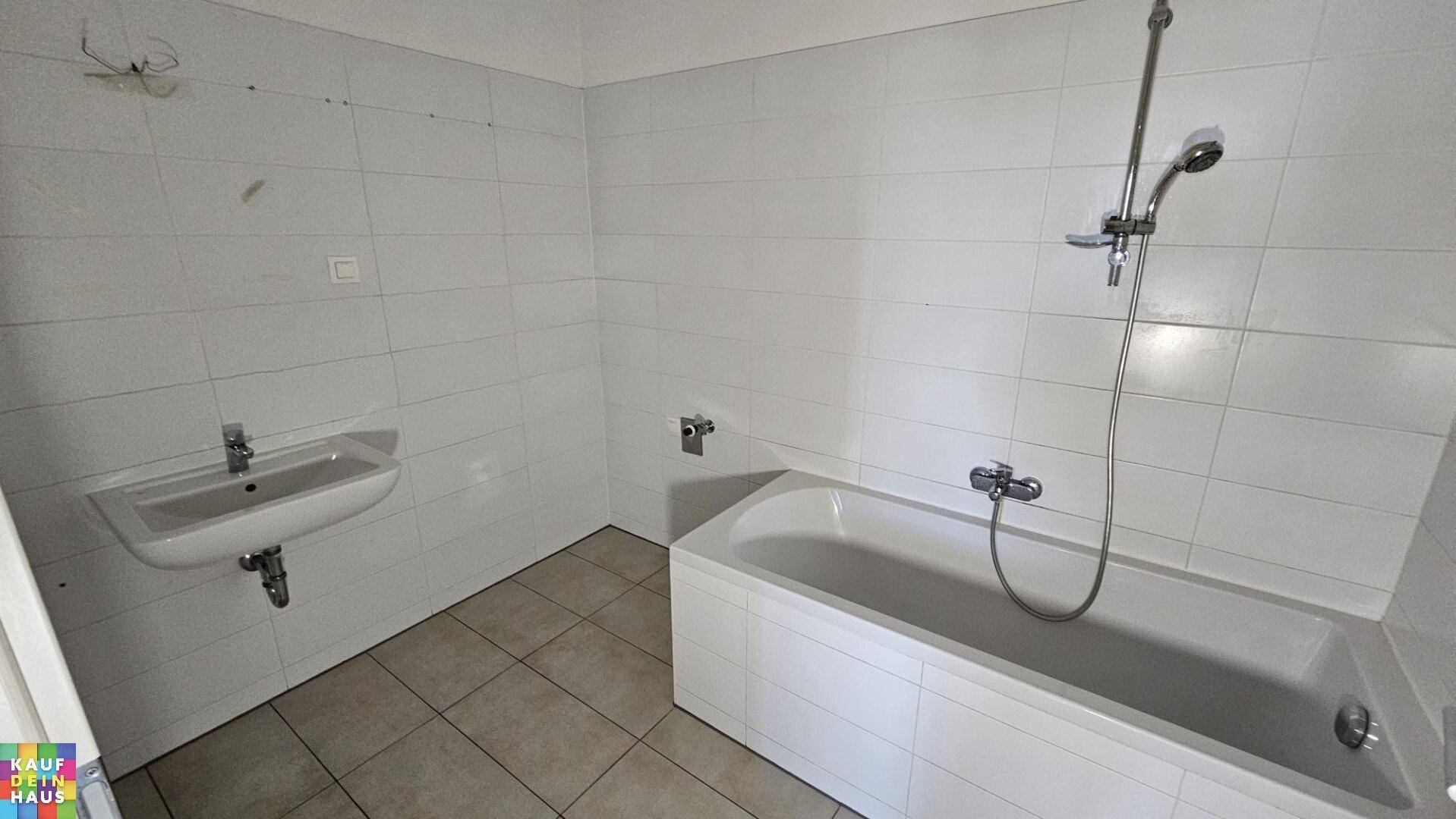 Badezimmer mit weißen Fliesen, Waschbecken und einer Badewanne mit Duschfunktion.