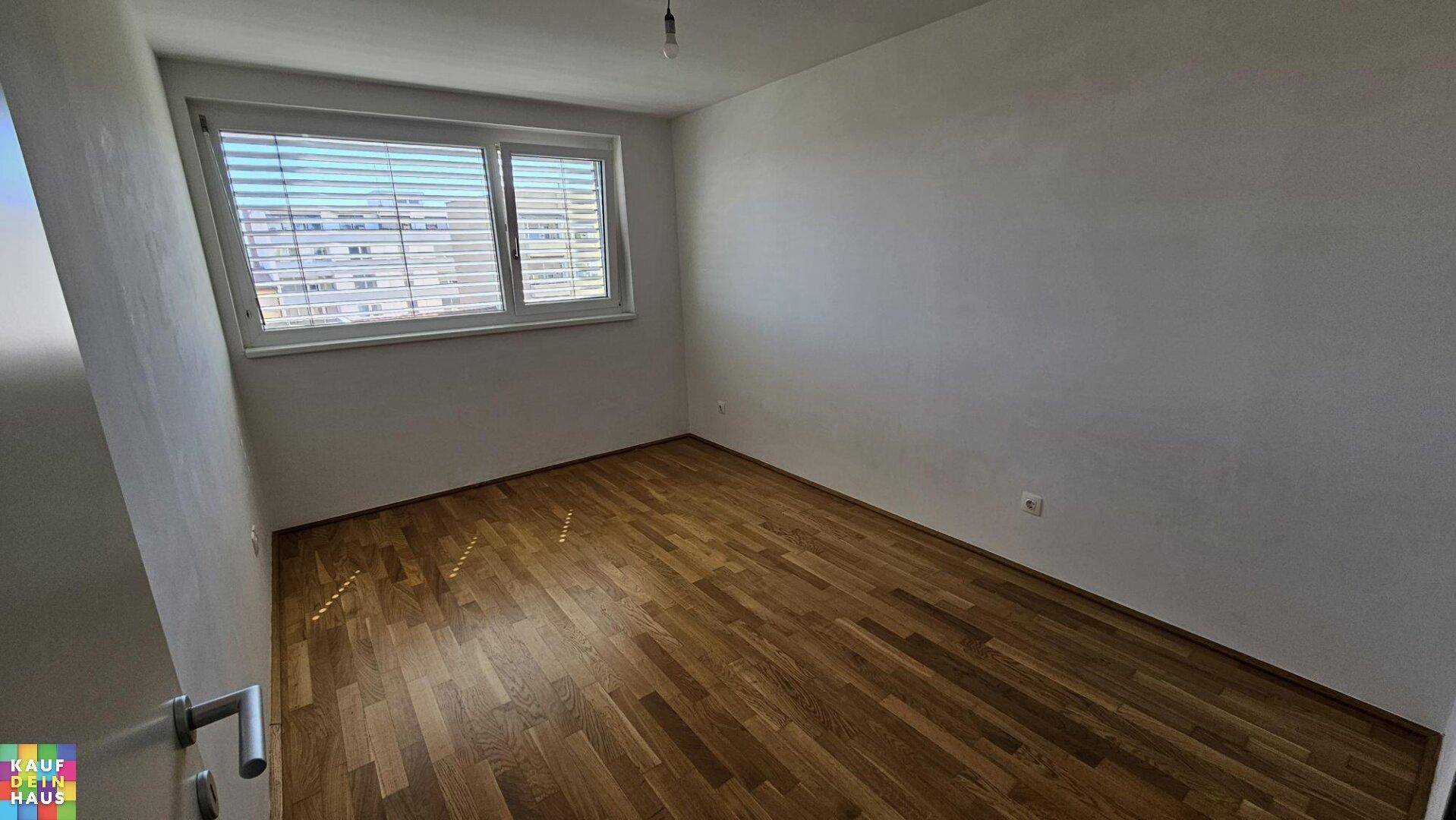 Leeres Schlafzimmer mit Parkettboden und einem Fenster, das viel Tageslicht hereinlässt.