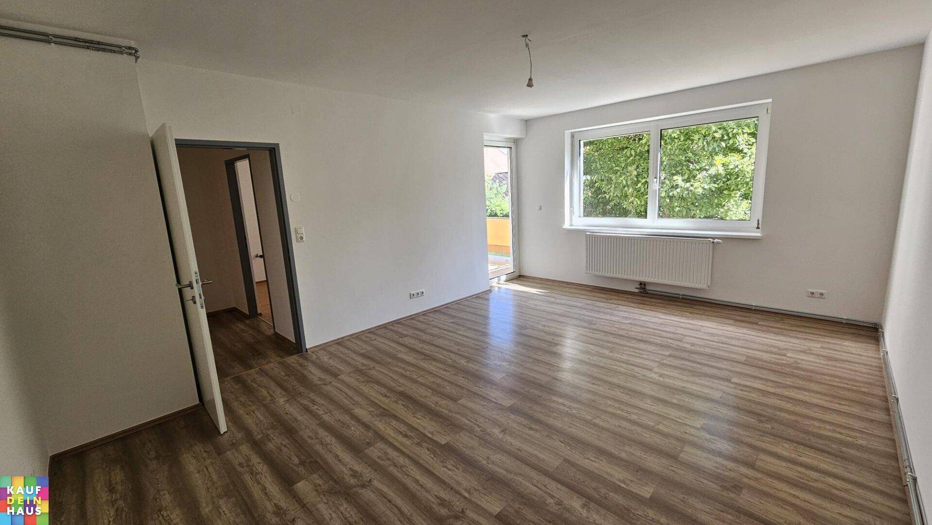 Helles, leeres Wohnzimmer mit modernem Laminatboden und Zugang zu einem Außenbereich.