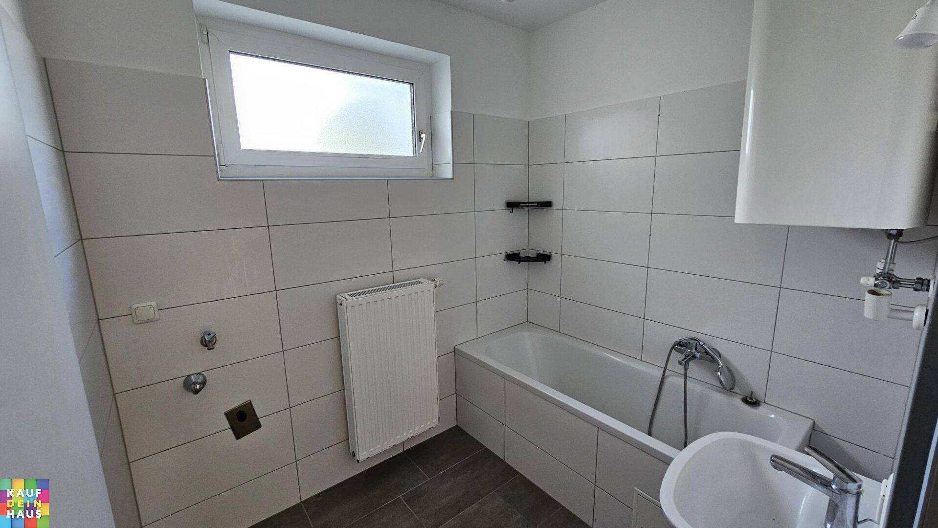 Funktionales Badezimmer mit weißen Fliesen, Badewanne, kleinem Waschbecken und Fenster.