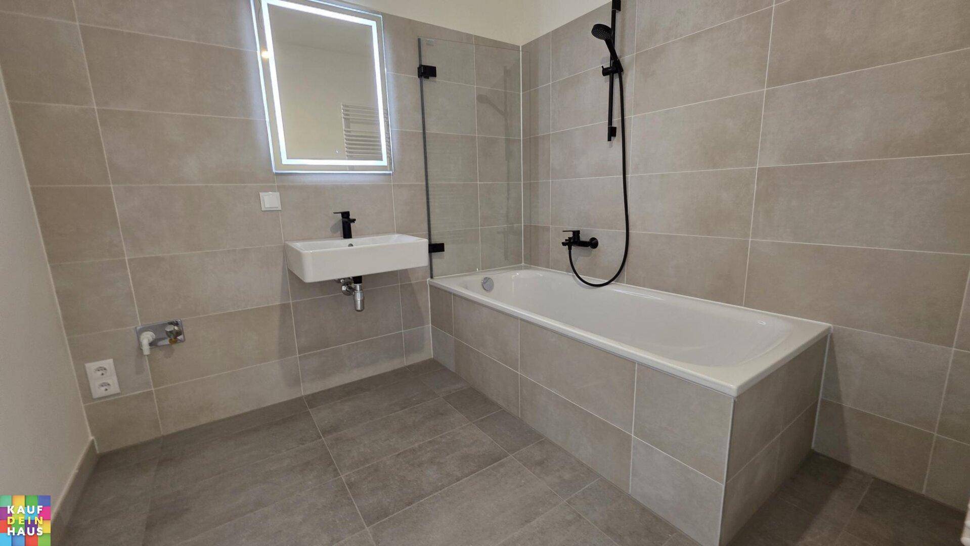 Modernes Badezimmer mit grauen Fliesen, Badewanne mit Duschfunktion und beleuchtetem Spiegel.