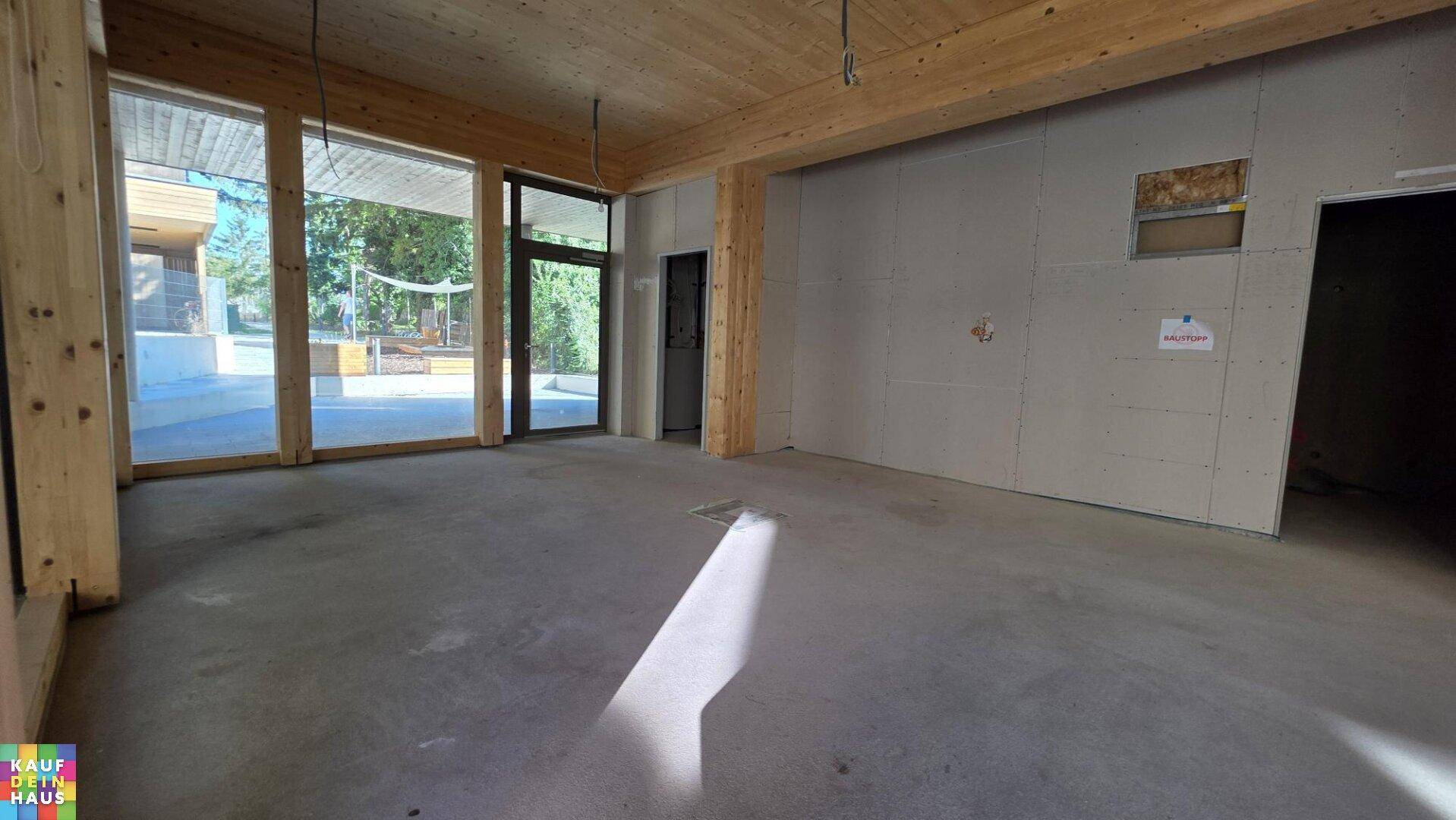 Großer, heller Innenraum im Rohbau mit Holzbalkendecke und bodentiefen Fenstern zur Außenseite.