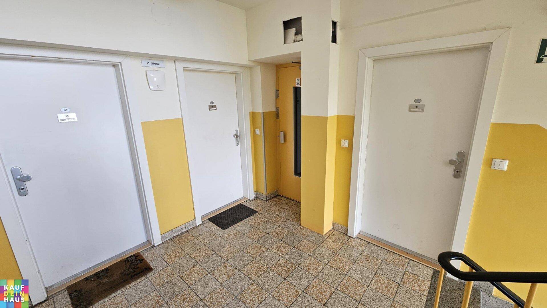 Gemeinschaftlicher Flur mit mehreren Wohnungstüren und einem Aufzug im Hintergrund.