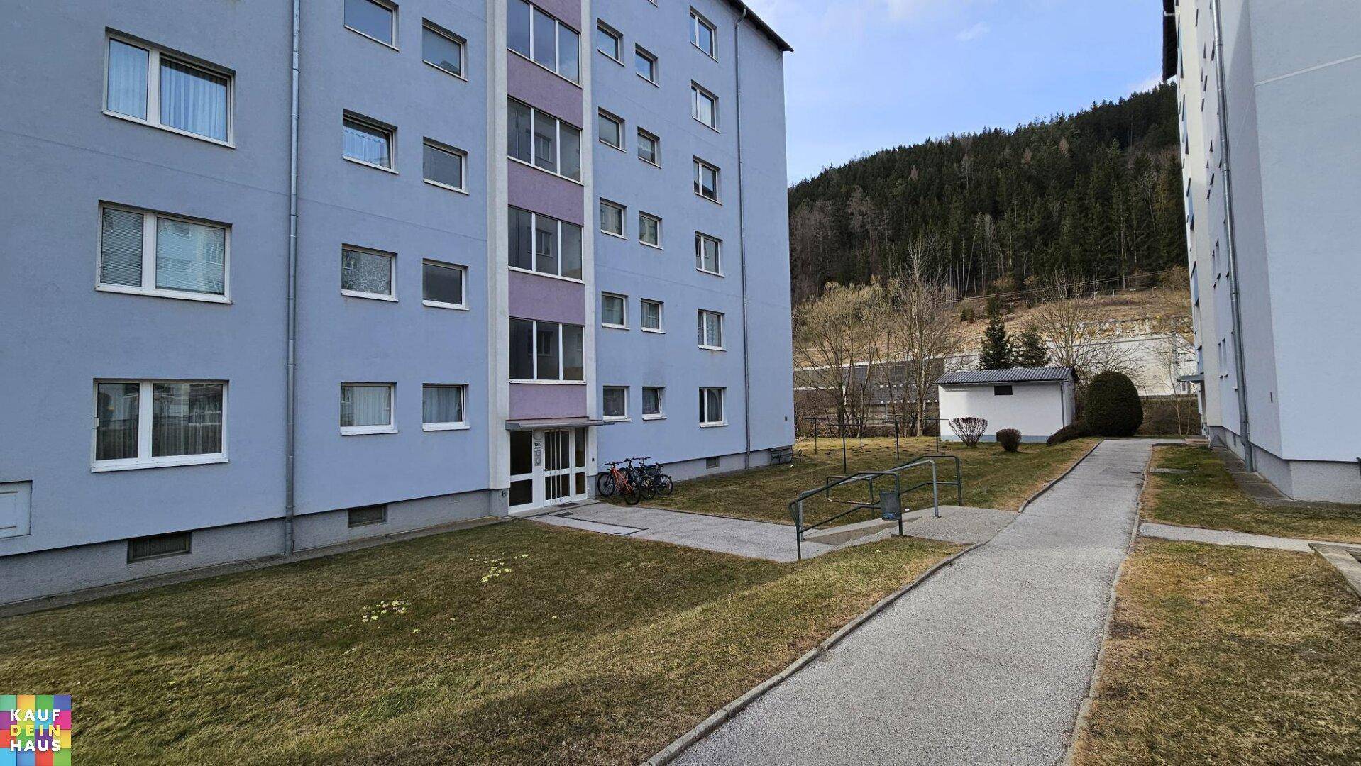 Wohngebäude mit blauer Fassade, umgeben von Rasenflächen und Bäumen, mit Hügel im Hintergrund.