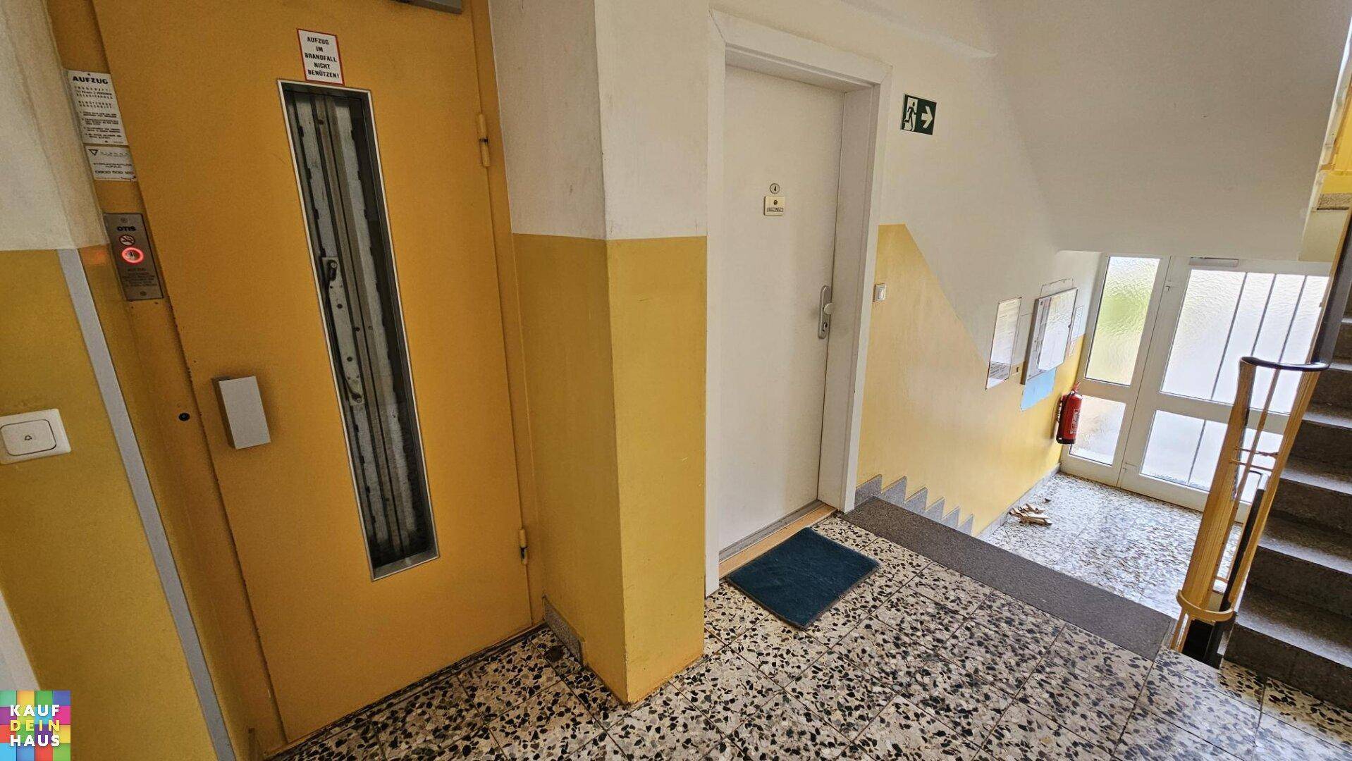 Eingangsbereich eines Mehrfamilienhauses mit Treppenhaus und gelbem Aufzug.